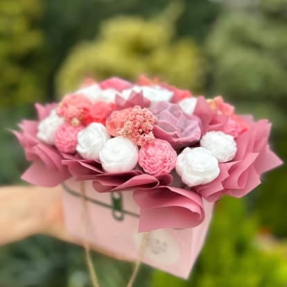Pink and white carnation flowers arranged in a pink box held by a person against a blurred green garden background