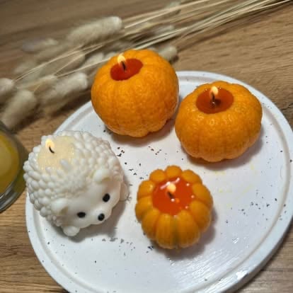 Plate with four decorative desserts: two large orange gulab jamun with red centers, a small white hedgehog-shaped pastry, and a small orange fluted cake, on white plate with dried grass in background