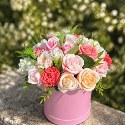 Pink flower arrangement in a pink cylindrical box with roses, carnations, and greenery displayed outdoors