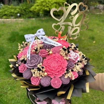 Decorative bouquet with pink and purple soap flowers, wooden heart topper, gold ribbon, and congratulations card held in garden