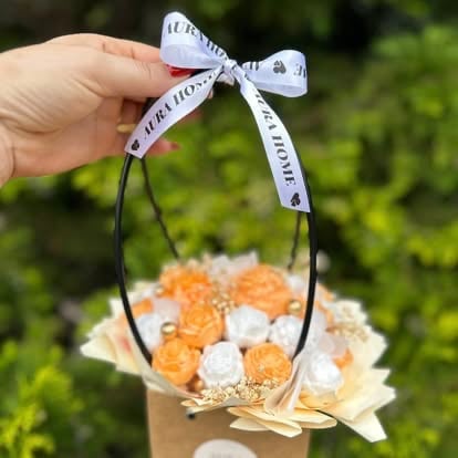 Hand holding a decorative gift basket with orange and white candies tied with a white ribbon bow against a blurred green background