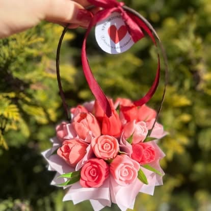 Hand holding a decorative floral arrangement with pink roses, ribbon bow, and circular ornament against blurred green foliage background