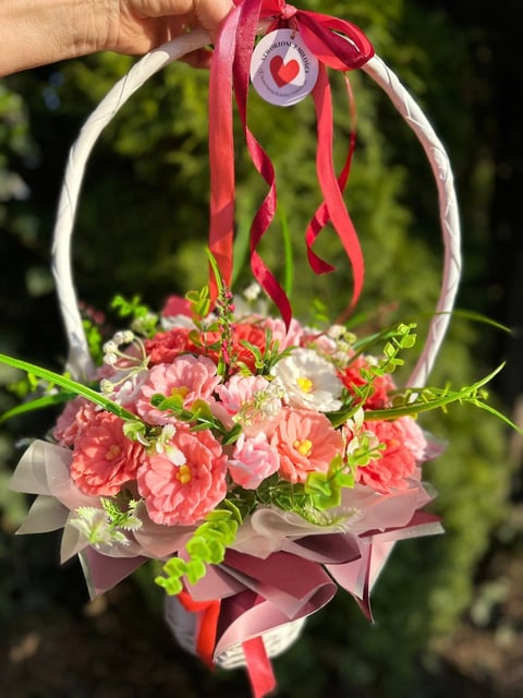 Hand holding a decorative flower basket with pink carnations, white handle, red ribbons, and a heart-shaped gift tag outdoors